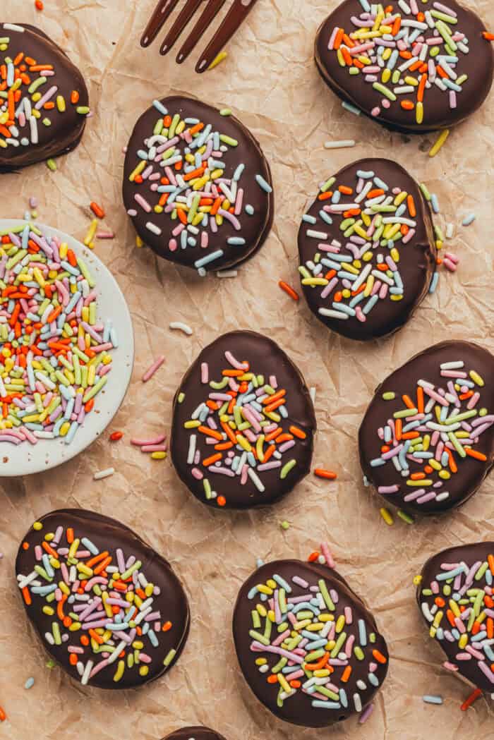A few homemade chocolate Easter eggs scattered on parchment paper with a small plate filled with sprinkles.