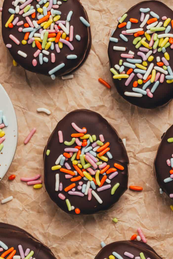 A few homemade chocolate Easter eggs scattered on parchment paper with a small plate filled with sprinkles.