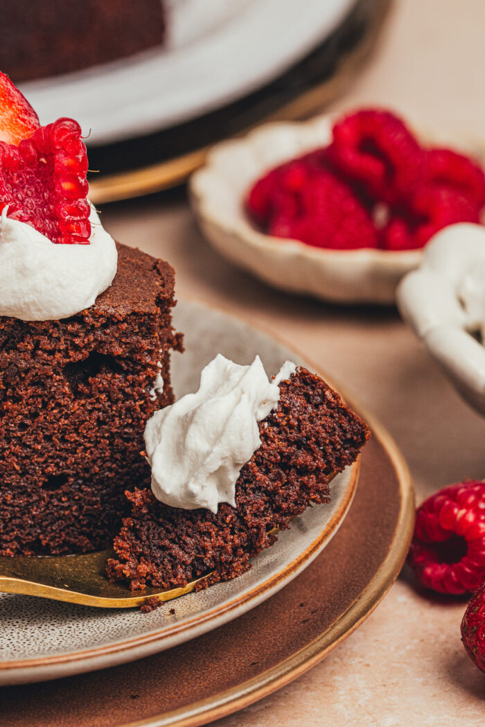 A slice of chocolate ricotta cake with whipped cream and berries with a bite taken out of it on a plate.