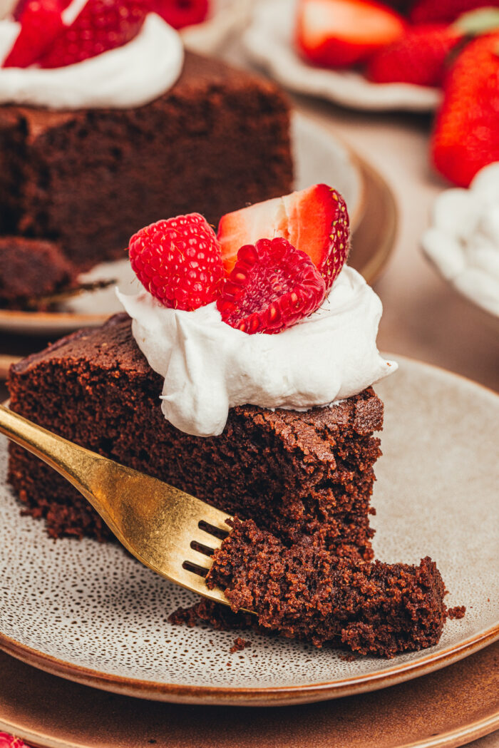 A slice of chocolate ricotta cake with whipped cream and berries with a bite taken out of it on a plate.
