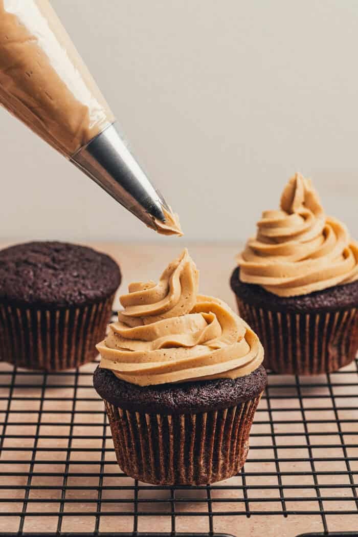 A piping bag with espresso frosting being piped onto the chocolate cupcakes.