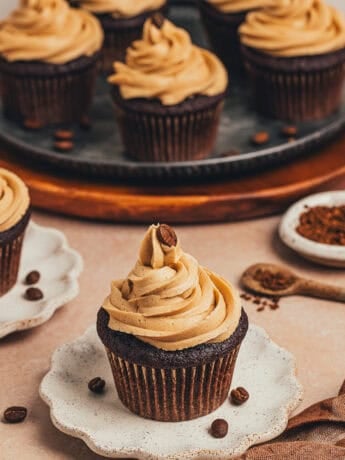 A chocolate cupcake with espresso buttercream frosting on a white plate with a platter of cupcakes behind it.