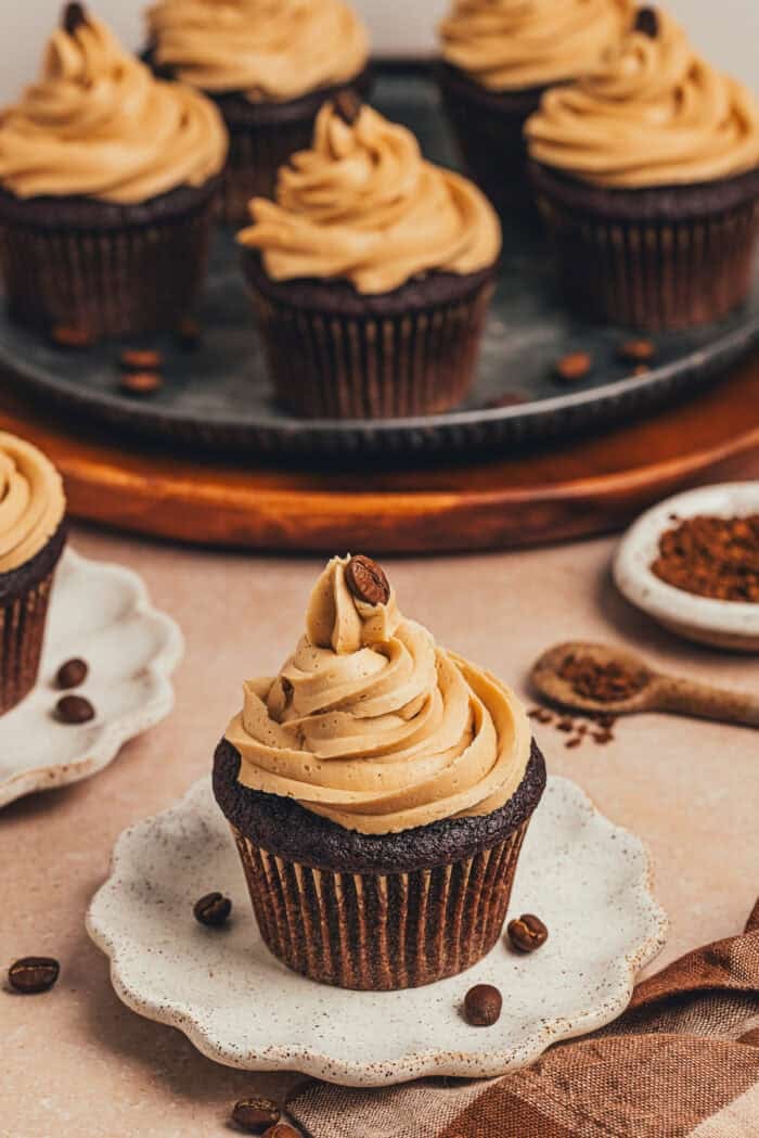 A chocolate cupcake with espresso buttercream frosting on a white plate with a platter of cupcakes behind it.