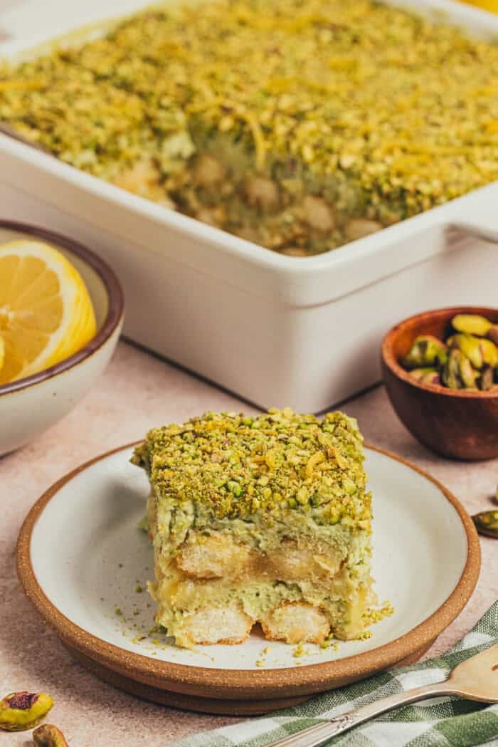 A slice of lemon pistachio tiramisu on a plate with a cake pan of tiramisu in the background.