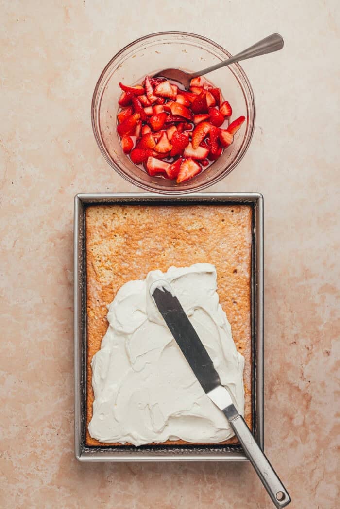 Whipped cream being spread over the tres leches cake base with a bowl of strawberries nearby.