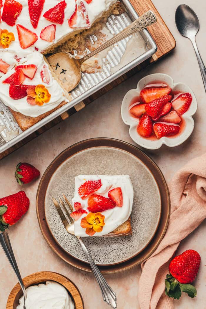 A slice of strawberry tres leches cake on a plate with a small bowl of fresh sliced strawberries.
