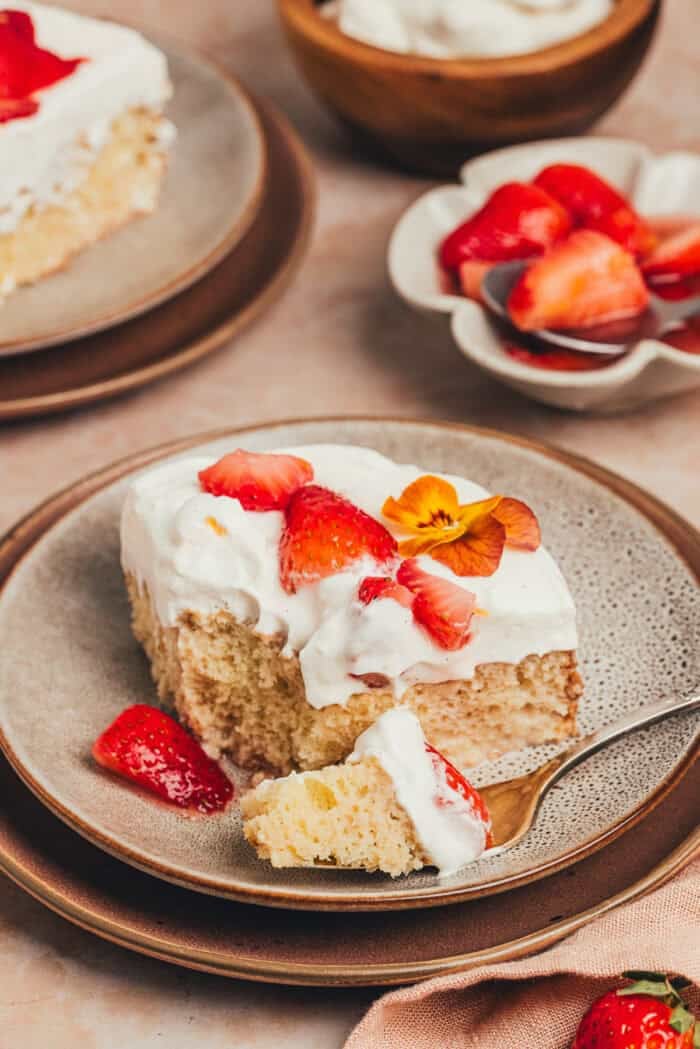 A slice of strawberry tres leches cake on a plate with a fork taking a bite out of the cake.