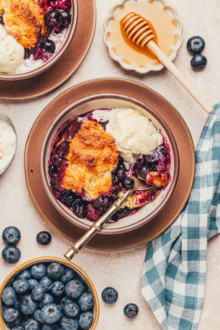 A bowl with a serving of blueberry cobbler and a spoon with various bowls of ingredients.