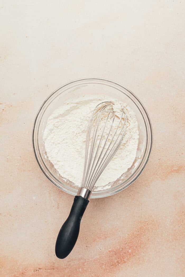 A glass bowl with flour and the other dry ingredients with a whisk.