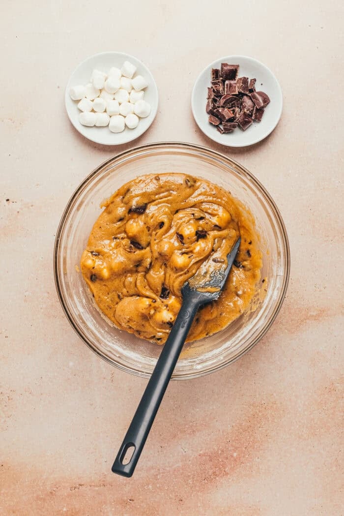 The s'mores bar batter in a glass bowl with a spatula and two small bowls with marshmallows and chocolate chips.