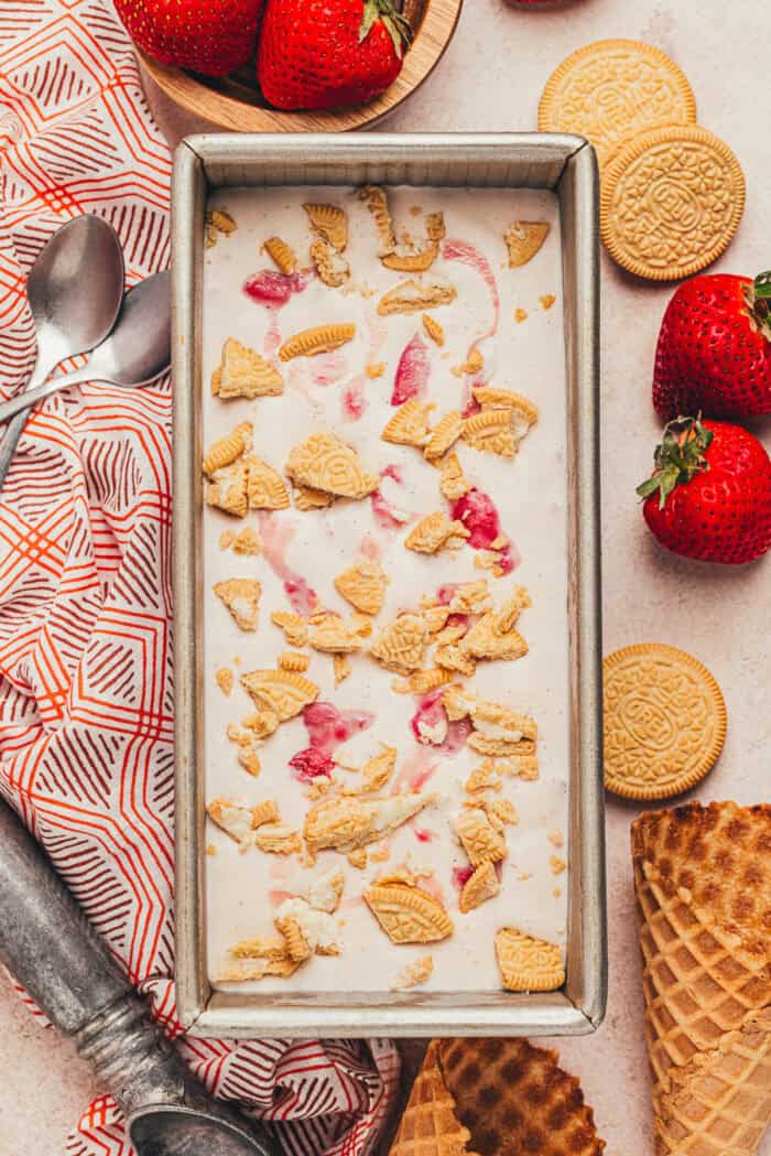 The overhead view of the top of the homemade no churn strawberry ice cream in a loaf pan with spoons and strawberries surrounding it.