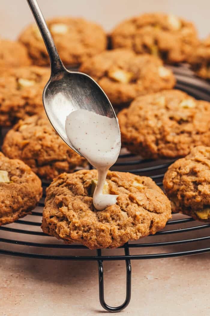 A spoon with icing is being drizzled over apple oatmeal cookies on a cooling tray.