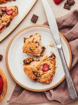 A chocolate strawberry scones on a white plate with chocolate chunks around it and fresh strawberries.