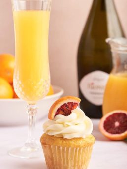 A mimosa cupcake with a glass of mimosa and a glass bottle of orange juice and Prosecco in the background.