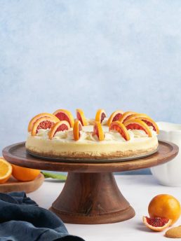 A blood orange cheesecake with sliced blood oranges on a wooden cake plate against a blue background.