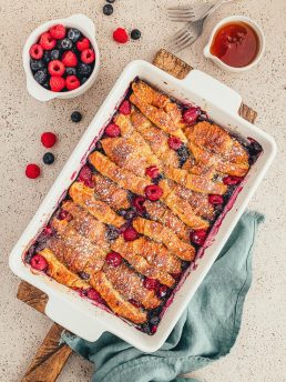 Croissant French toast bake in a white casserole dish with a bowl of berries and a teal cloth napkin.