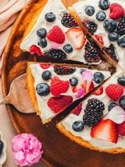 A beautiful berries and cream cake on a wooden platter that has been cut into slices.