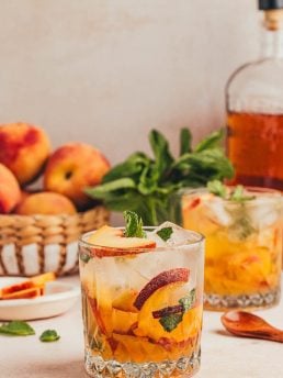 A glass filled with a bourbon peach smash with a bowl of peaches in the background and a bottle of bourbon.