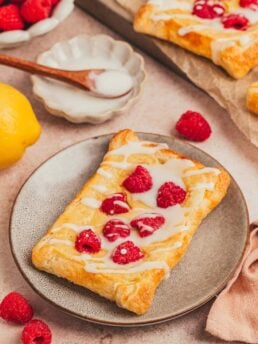 A homemade raspberry lemon cream cheese danish on a plate with a small bowl of icing with a spoon.